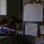 calendário de papel preso na geladeira com dias marcados em cores diferentes, ao lado de um horário escolar e uma mochila infantil
