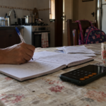 mão de mulher escrevendo contas em um caderno sobre a mesa da cozinha, com calculadora de celular ao lado e mochila escolar ao fundo